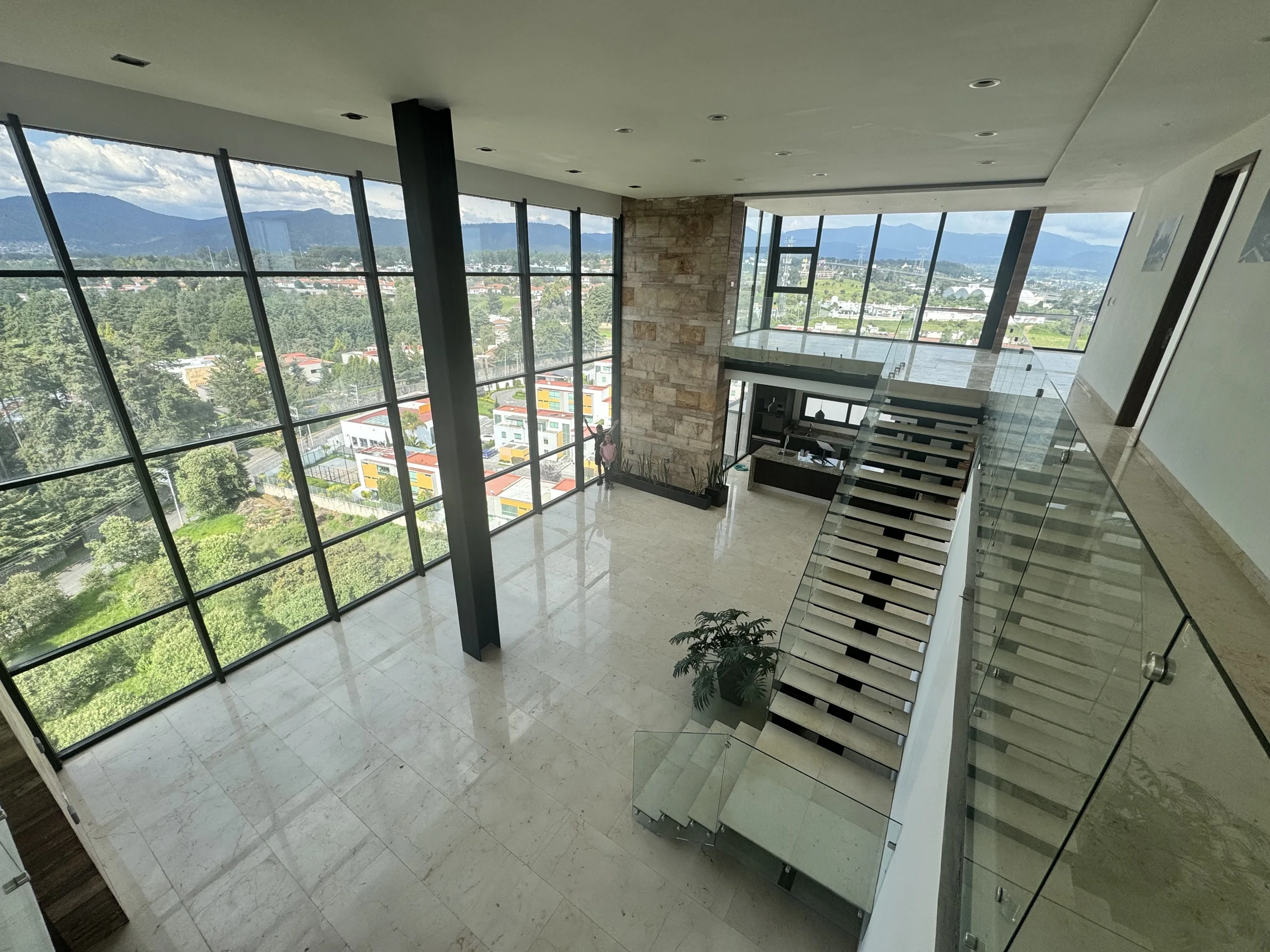 Doble altura con ventanal de piso a techo y escalera flotante de mármol y cristal, con vista al bosque y las montañas del valle de Lerma