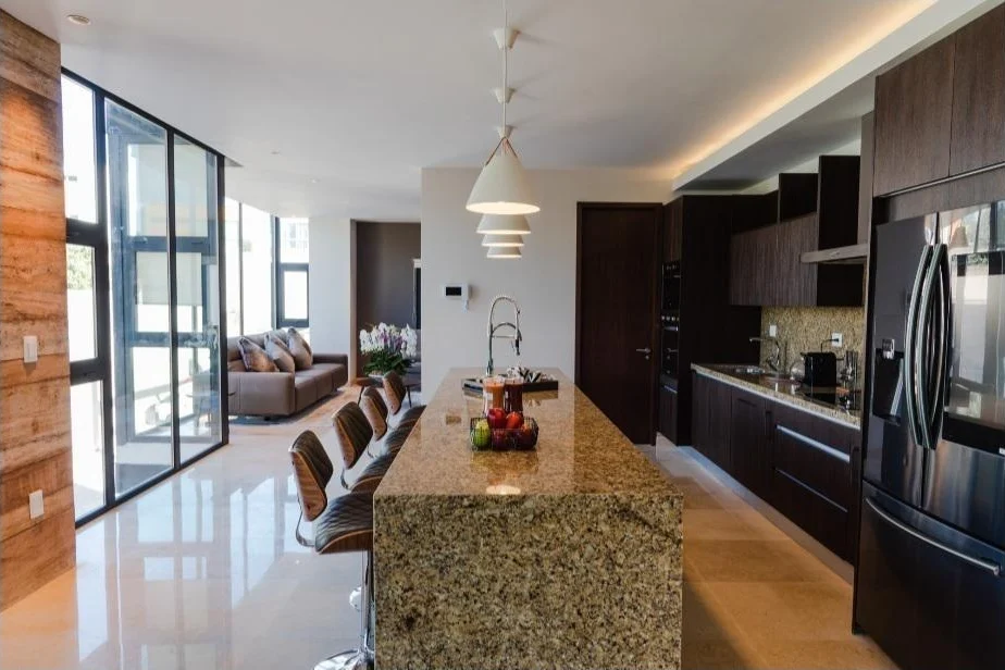 Cocina con isla de granito abierta hacia la sala, con ventanal de piso a techo